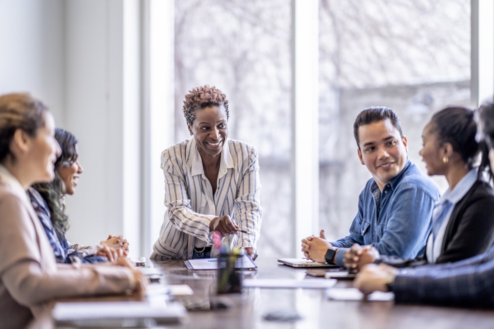 A small group of business professionals sit around a table as they meet to brainstorm some ideas for the future of the company. They are each dressed professionally and have papers scattered out in front of them as they work together.