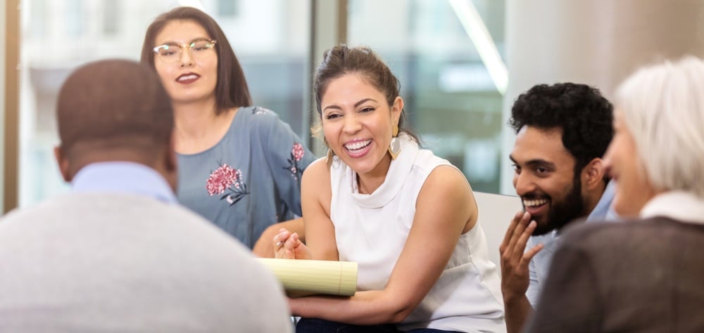 A young businesswoman sits in a circle with a group of colleagues. She holds a note pad as she laughs with them.