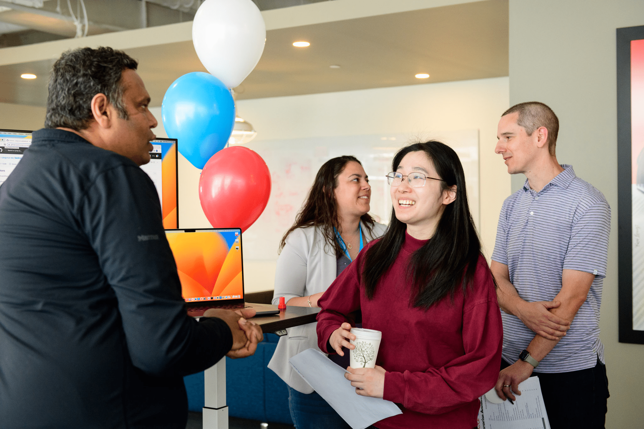 Man and woman engaging in conversation at a CarGurus event. They are next to a table with a computer on it and three balloons - red, blue and white. There are two people behind them also chatting.