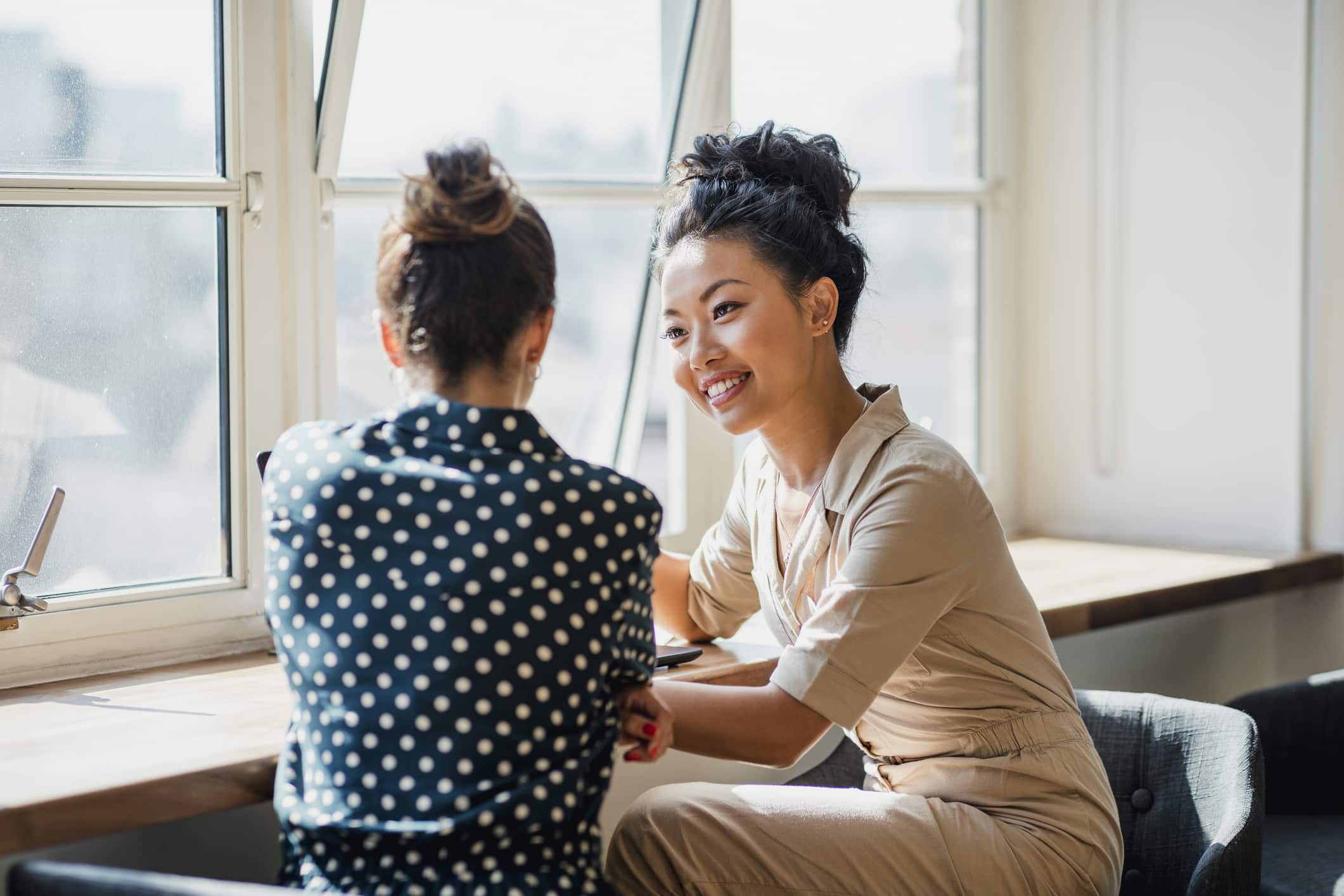 Two women colleagues laughing while standing near a window together at their workplace. There is a laptop on the table in front of them