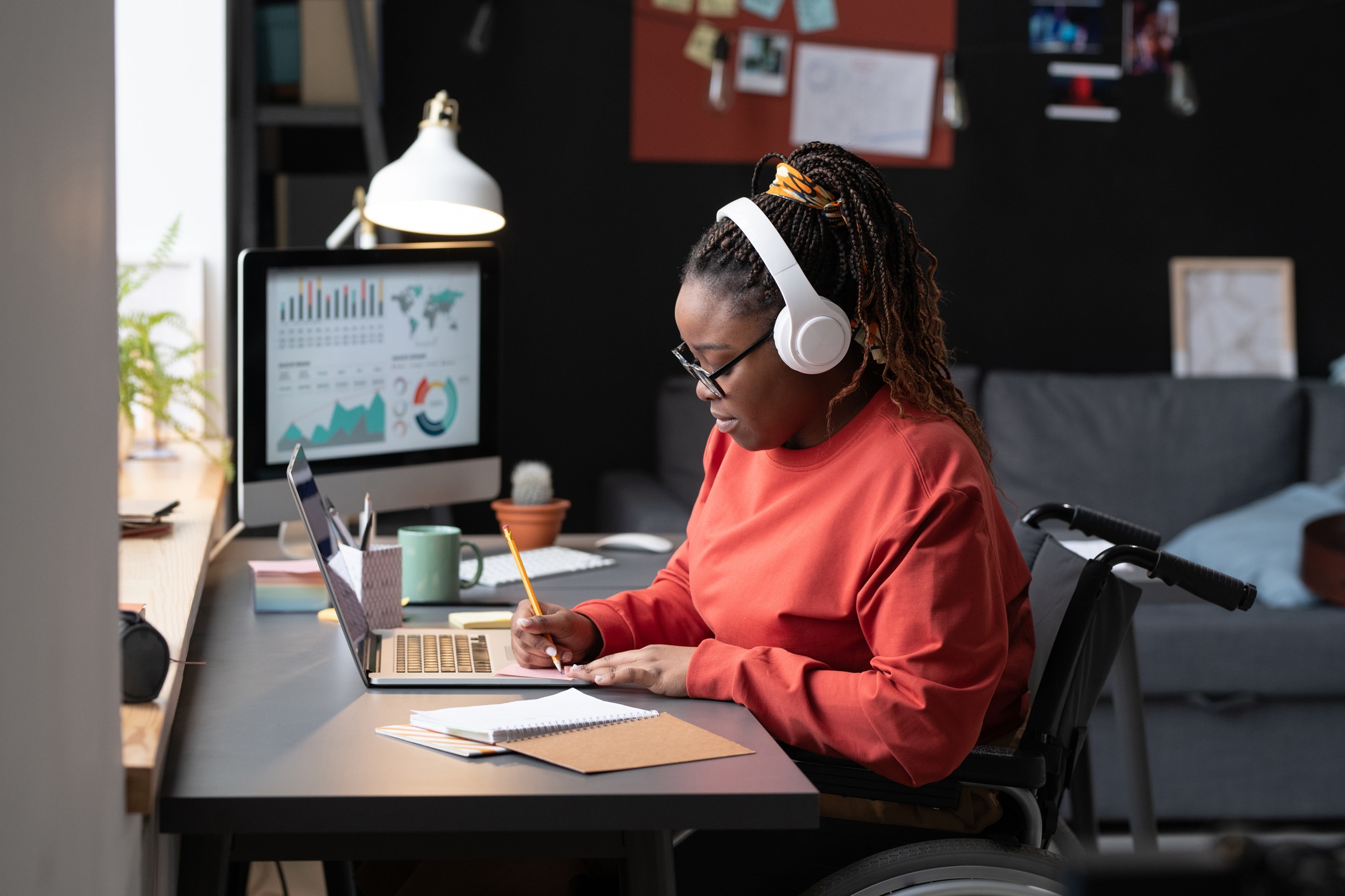 Black woman in wireless headphones sitting at the table in front of the laptop and making notes, she studying at home.