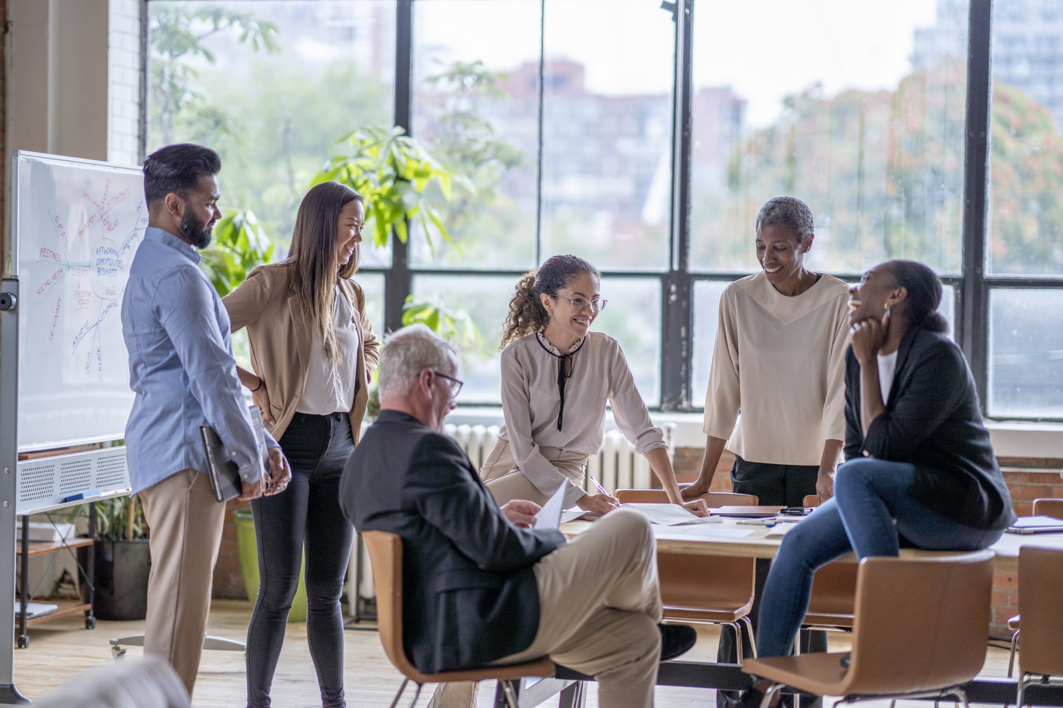 A small group of business professionals casually meet in a downtown office as they discuss new strategies and goals for the company. They are each dressed professionally and are casually sitting around and on a boardroom table.