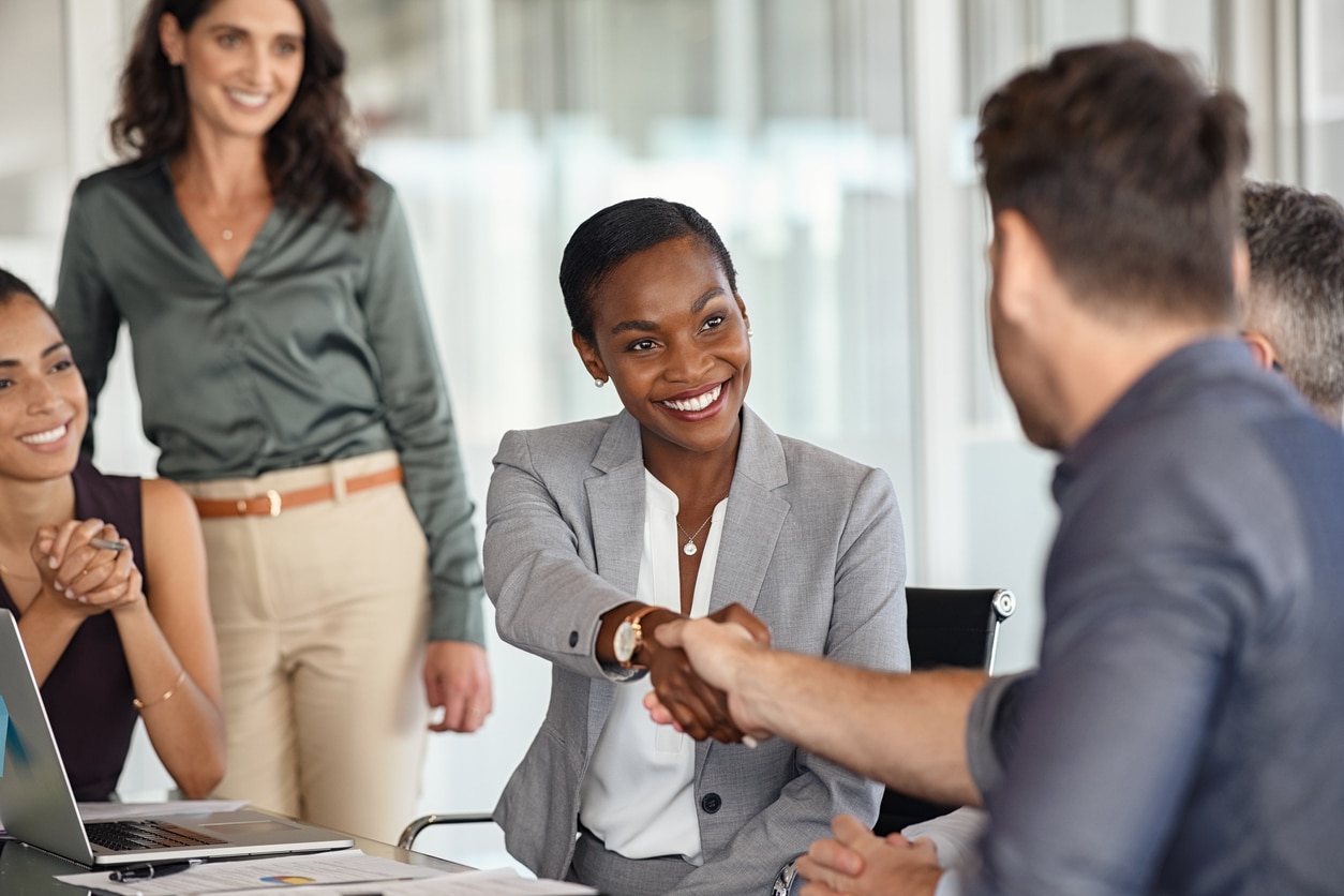 Black professional woman shaking hands with a coworkers at meeting and other employees sitting and standing in the background.