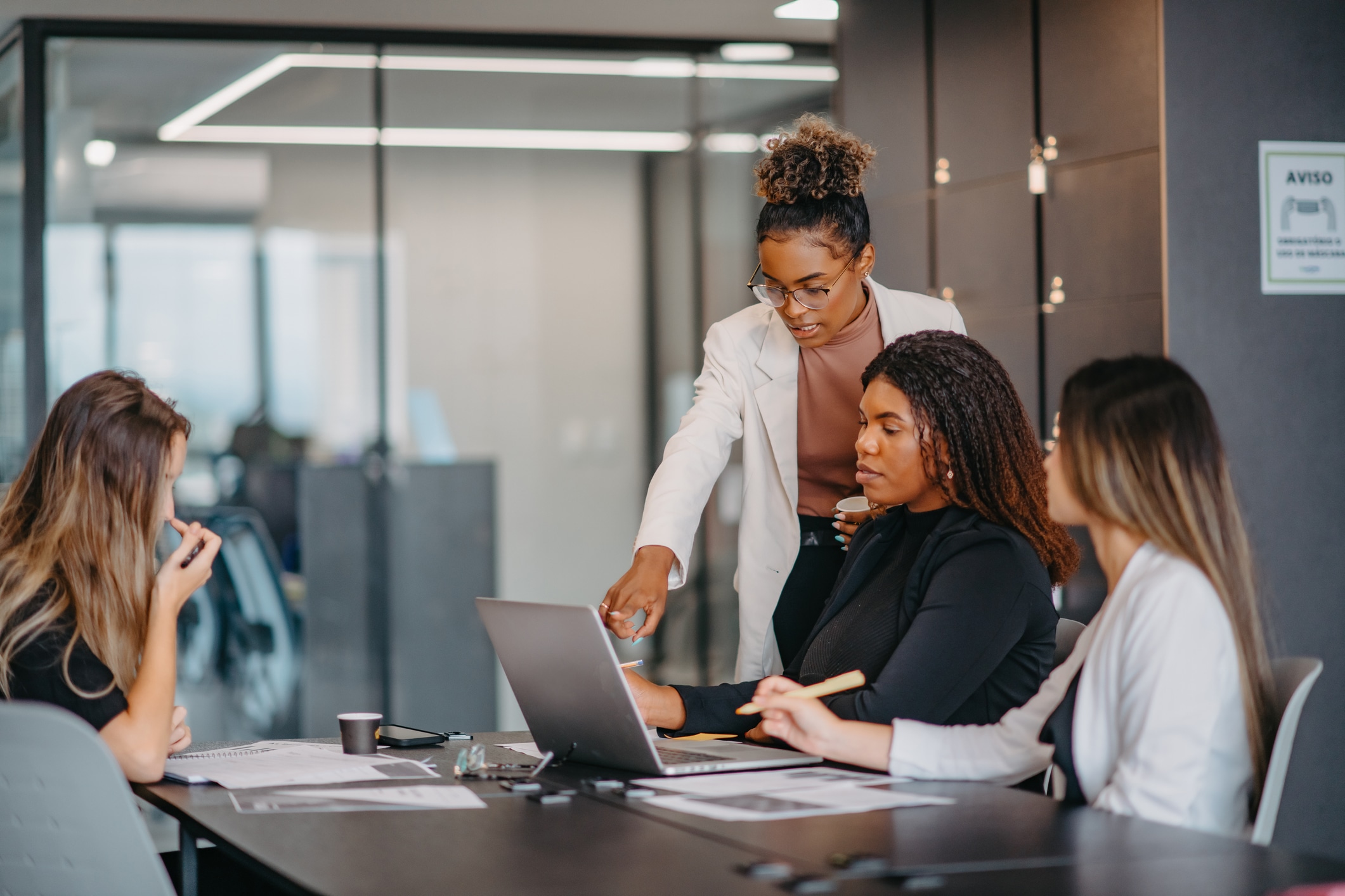 A group of women during a meeting in the office. One women is standing and pointing to something at the laptop on the table.