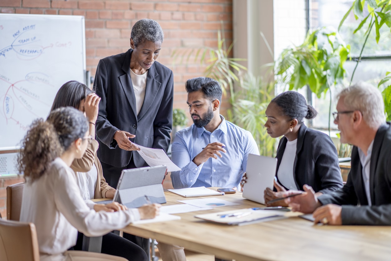 A small group of mature corporate business men and women, gathered around a boardroom table as they meet to discuss the company.