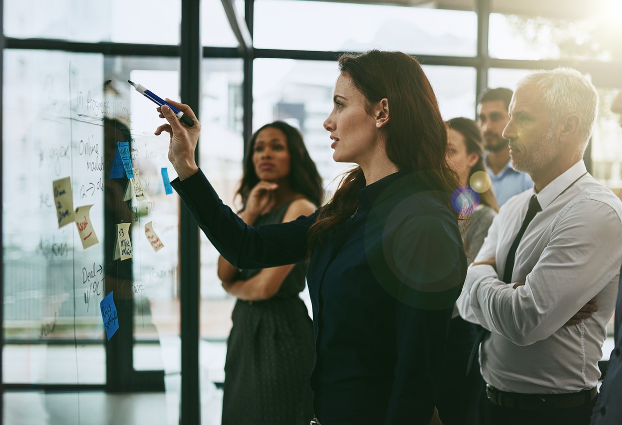 Businesswoman writing on a glass window and strategizing with a group of business colleagues behind her during a meeting in the boardroom.