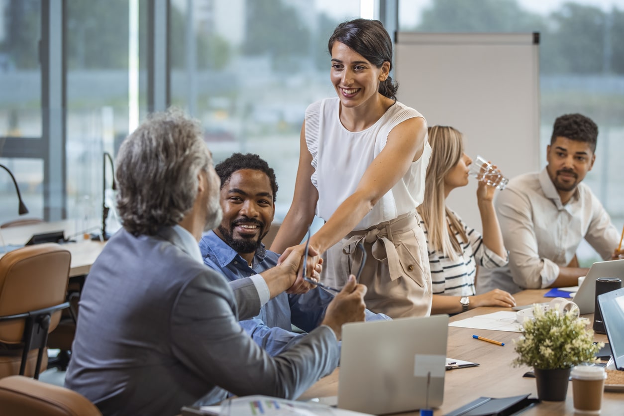 Shot of a seated man and a standing woman shaking hands during a boardroom meeting. Colleagues are also gathered at the table.