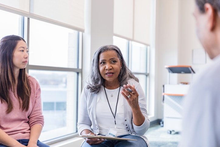 In a group meeting, a young woman listens as the senior adult female leader gives feedback to an unrecognizable man.