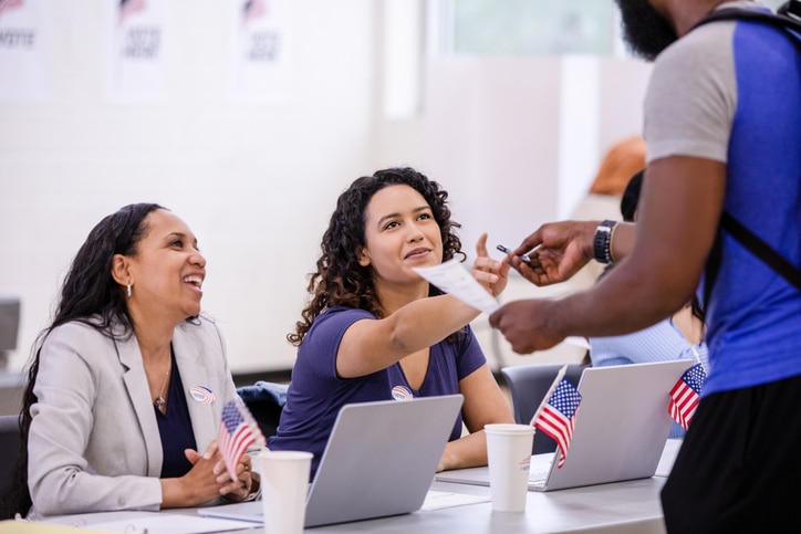 Two cheerful female volunteers help a young adult with his ballot for the election.