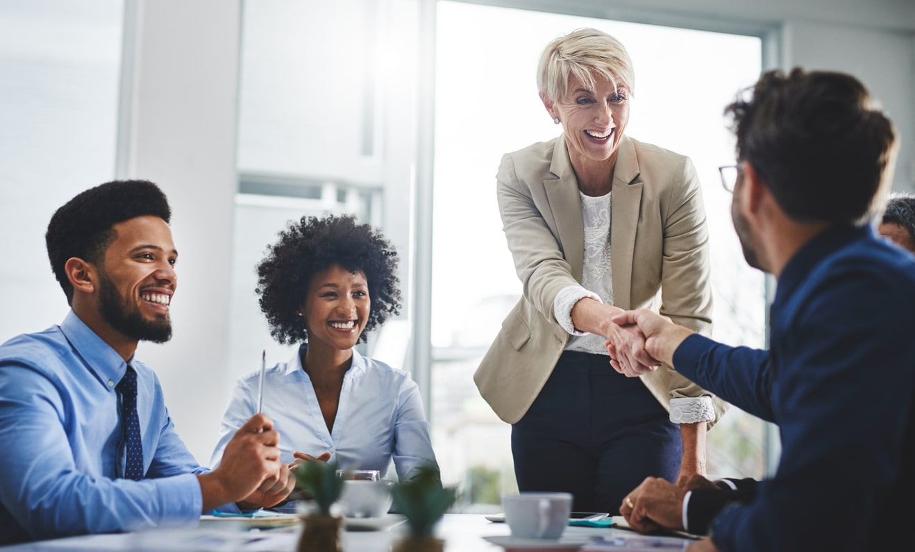 Standing business woman shaking hands with a seated business man with his back to the camera. They are in a team meeting at the office.