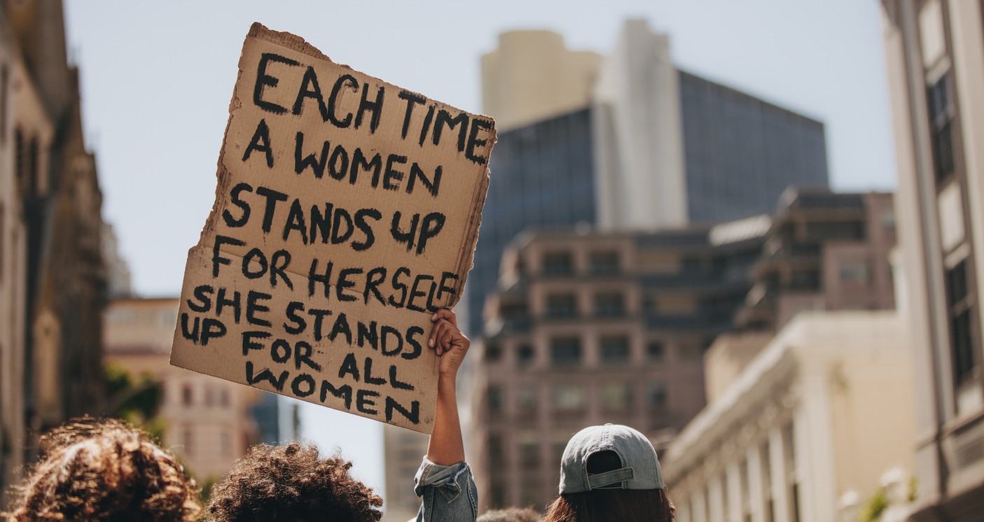 Group of demonstrators on road with a cardboard sign that reads 'Each time a woman stands up for herself she stands up for all women,' in all caps.