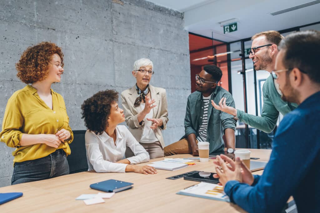 A group of businesspeople gathered together in a meeting. Two are sitting, three are standing, and one is sitting on the edge of the table.