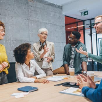 A group of businesspeople gathered together in a meeting. Two are sitting, three are standing, and one is sitting on the edge of the table.