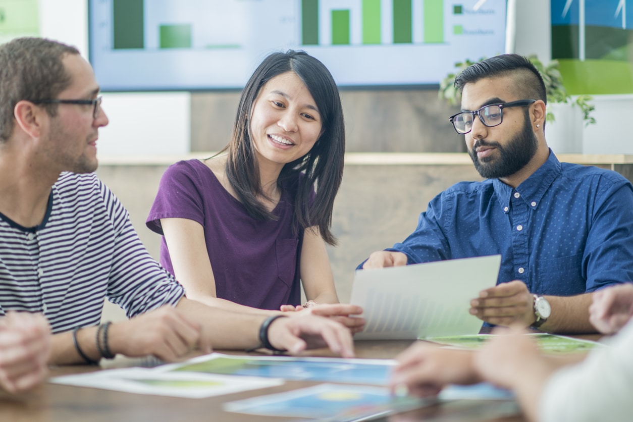 A group of business-people are indoors in a board room. They are wearing casual and semi-casual clothing. They are discussing plans for a green energy project. Many chart and graphs are on the table.